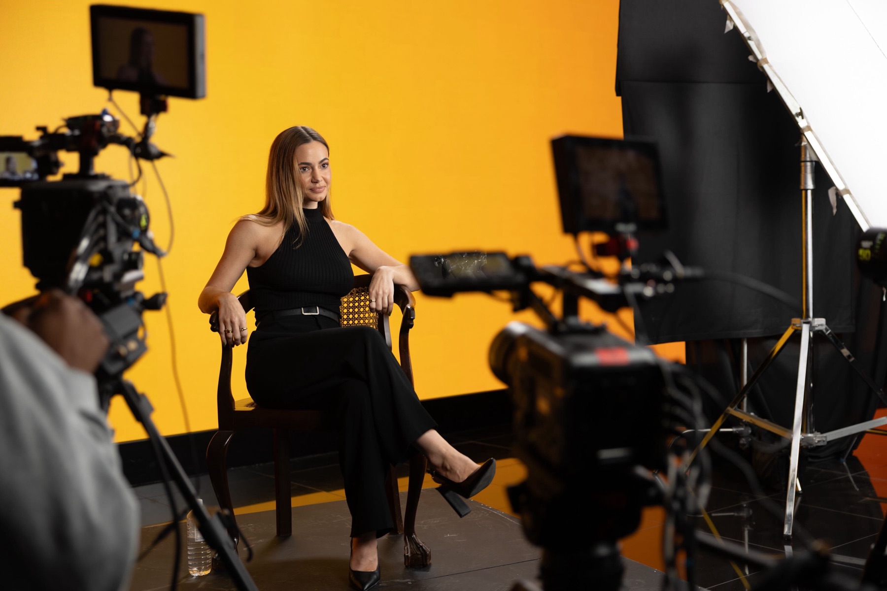 Kelly on a studio set, seated in front of cameras against a yellow backdrop
